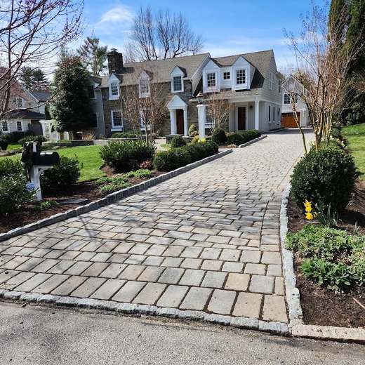 A cobblestone-style paver driveway edged in granite cobble, running up to a stone and clapboard house between freshly mulched beds.